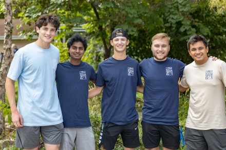 Five young men standing arm in arm and smiling at the camera outside